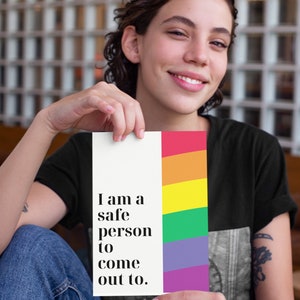 May include: A person holding a white card with the text "I am a safe person to come out to." The card has a rainbow stripe on the right side.