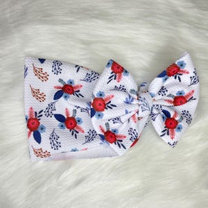 May include: White headband with a large red, blue, and white floral bow. The headband is made of a soft, stretchy fabric and is perfect for adding a touch of style to any outfit.