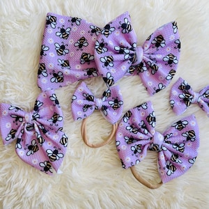 May include: Three purple and white bee-themed hair bows with matching headbands. The bows are made of soft fabric and have a cute bee design. The headbands are elastic and fit comfortably on a child's head.
