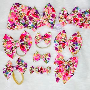 May include: A set of 8 floral print hair bows in various sizes. The bows are made of pink, white, and green fabric with a floral pattern. The bows are tied with a matching elastic band.