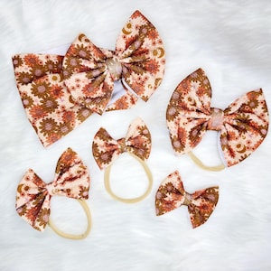 May include: A collection of five hair accessories featuring a floral and celestial pattern in shades of orange, brown, and cream. The set includes one wide headband with a large bow, three smaller bows on thin elastic bands, and one small bow clip.