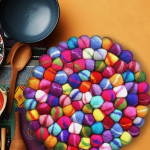 Puede incluir: Un posavasos redondo y colorido hecho de bolas de lana fieltro. Las bolas están dispuestas en espiral y son de tonos rosa, morado, azul, verde, amarillo, naranja y rojo.