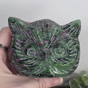 May include: A polished stone carving in the shape of an owl head, featuring a mottled pattern of deep forest green and vibrant magenta-pink mineral inclusions. The sculpture displays carved details for the eyes, beak, and feathered facial features.