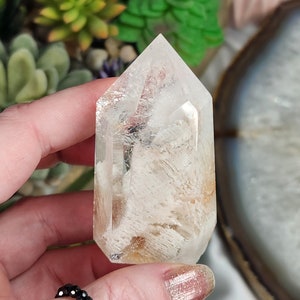 May include: A clear quartz crystal point with a white and gold inclusion. The crystal is pointed at the top and has a flat base.