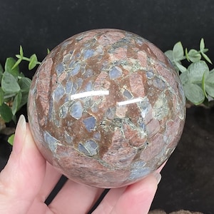 Peut inclure: Une pierre sphérique polie présentant une mosaïque d'inclusions minérales brun rougeâtre, bleues et grises. La surface est lisse et réfléchissante, affichant un motif complexe de formes cristallines imbriquées sur toute la sphère.