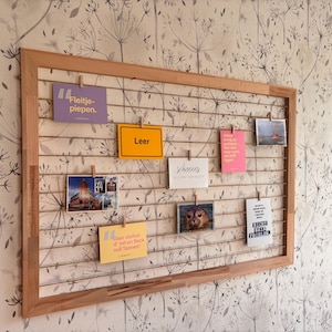 Op de afbeelding: Een houten wanddecoratie met een raster waarop verschillende ansichtkaarten en notities met wasknijpers zijn bevestigd. De ansichtkaarten bevatten afbeeldingen en tekst in het Nederlands, waaronder "Leer", "schvenes aus der Werkdat", "Daar steihst d' mit en Beck vull Tannen!" en "NICHT MEIN PROBLEM".