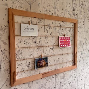 Op de afbeelding: Een houten displayframe met touwlijnen voor het ophangen van foto's en notities. Het frame bevat drie kaarten: één met tekst, één met schapenillustraties en een foto van een zeehond. De achtergrond is een behang met bloemenpatroon.