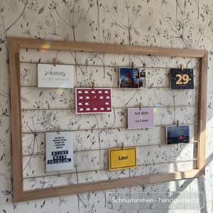Peut inclure: Un cadre d'affichage en bois avec plusieurs cartes postales et notes fixées à des cordes. Les cartes présentent divers motifs et textes, dont "schoenes", "NICHT MEIN PROBLEM" et "Leer". Le cadre est posé sur un fond de papier peint floral.