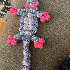 Peut inclure: Un jouet en peluche en crochet en forme de lézard avec une longue queue. Le lézard est violet, rose et bleu avec une tache blanche sur le dos.