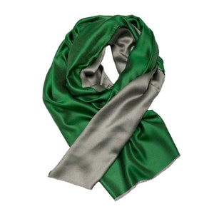 Puede incluir: Una bufanda reversible verde y gris. La bufanda está hecha de un material suave y sedoso y tiene un borde con flecos.