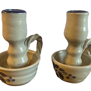May include: A pair of off-white ceramic candle holders with a speckled finish. Each holder features a circular base with a blue floral pattern, a central cylindrical candle socket with a blue rim, and a twisted handle attached to the side.