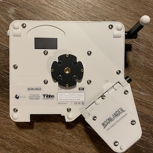 Puede incluir: Un dispositivo de pl&aacute;stico blanco con un centro negro y un m&oacute;dulo de pulgar blanco. El dispositivo est&aacute; etiquetado como "MOONLANDER" y "The Original Thumb Module".