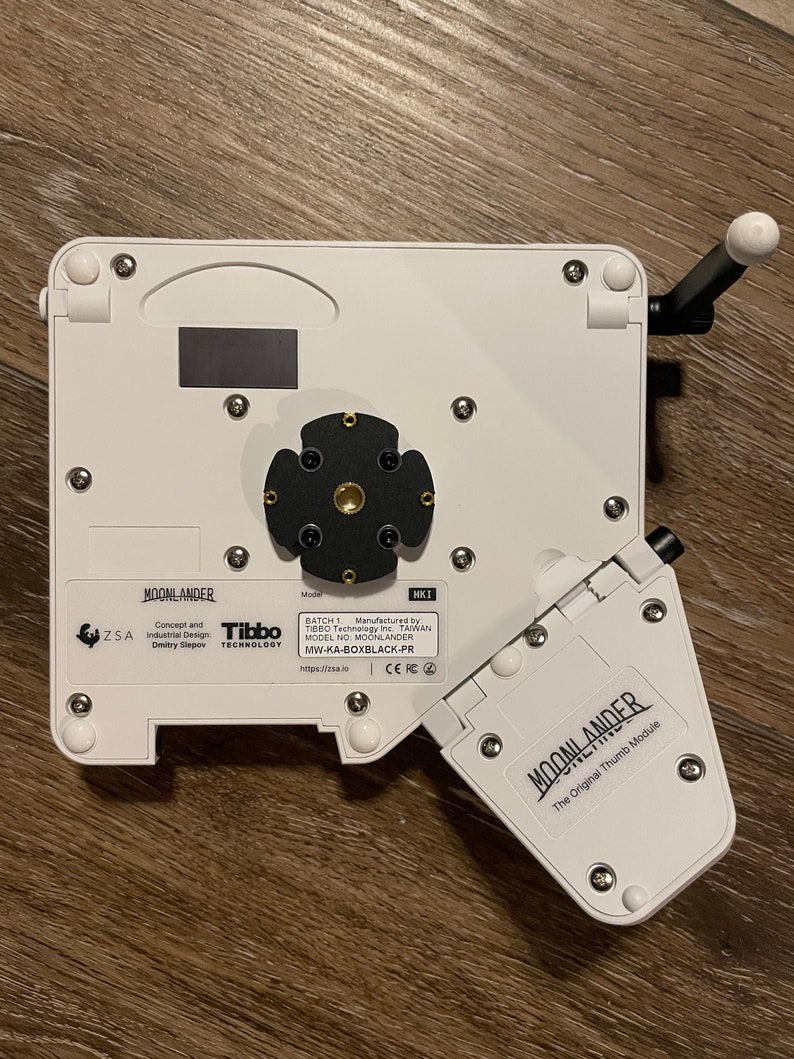 Puede incluir: Un dispositivo de pl&aacute;stico blanco con un centro negro y un m&oacute;dulo de pulgar blanco. El dispositivo est&aacute; etiquetado como "MOONLANDER" y "The Original Thumb Module".