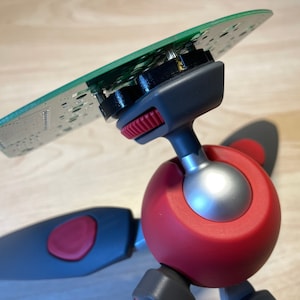 Puede incluir: Un mini tr&iacute;pode rojo y gris con una cabeza de bola y una placa de circuito verde unida a la parte superior.