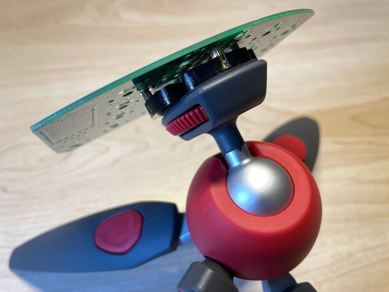 Puede incluir: Un mini tr&iacute;pode rojo y gris con una cabeza de bola y una placa de circuito verde unida a la parte superior.