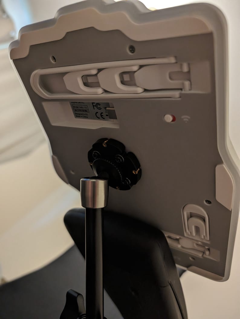 Puede incluir: Soporte de tableta blanco con un soporte giratorio negro y un brazo de metal negro. El soporte tiene un pestillo de pl&aacute;stico blanco en el lateral y una peque&ntilde;a luz roja en la parte posterior. El soporte est&aacute; unido a un brazo de metal negro que est&aacute; conectado a una base negra.