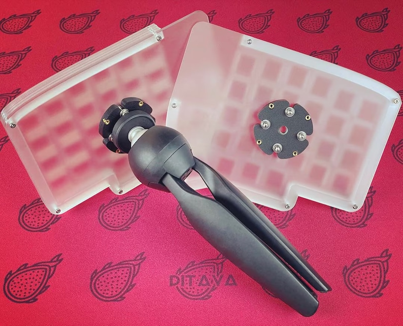 Puede incluir: Una carcasa de teclado de pl&aacute;stico transparente con un soporte de tr&iacute;pode negro y una placa circular negra con seis agujeros. La carcasa est&aacute; sobre una superficie roja con un patr&oacute;n de fruta de drag&oacute;n negra.