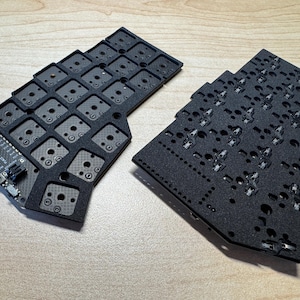 May include: Two black split mechanical keyboard plates with a grid of square cutouts. The left plate features a visible circuit board with a micro-USB port and printed text, while the right plate shows exposed metal switch sockets.
