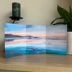 May include: A triptych artwork showing a tranquil beach scene at sunset. The painting uses blues, pinks, and oranges, with small figures on the water. The piece is displayed on a wooden surface.