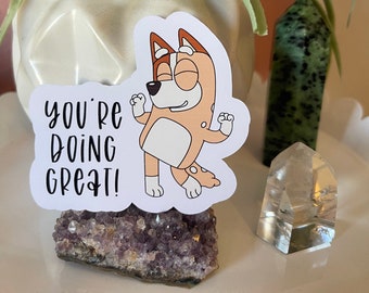 May include: A sticker featuring a cartoon dog with the text "YOU'RE DOING GREAT!" The dog is light brown, white, and blue. The sticker is placed on a purple amethyst crystal. A green plant and other crystals are in the background.