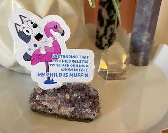 May include: A sticker featuring Bluey riding a pink flamingo, with the text "PRETENDING THAT MY CHILD RELATES TO BLUEY OR BINGO, WHEN IN FACT, MY CHILD IS MUFFIN." The sticker is on a white decorative plate with an amethyst geode.