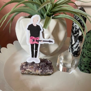 May include: A sticker of a person with blonde hair wearing a black shirt and ripped black jeans, holding a pink guitar. The sticker is placed on a purple amethyst crystal. A clear quartz crystal and a white geometric planter with a plant are also visible.