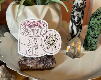 May include: A white sticker with the words "GRIND ON ME" and a cannabis leaf design. The sticker is on a purple amethyst crystal, displayed on a white decorative tray with other crystals and a potted plant.