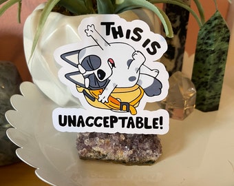 May include: A white sticker with a cartoon dog character, wearing a yellow bag, with the text "THIS IS UNACCEPTABLE!" The sticker is placed on a white decorative plate, with a purple crystal and a plant in the background.