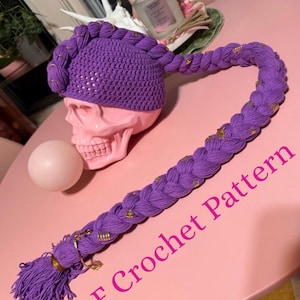 May include: A purple crochet hat with a long, braided extension, adorned with gold accents. The hat is resting on a pink skull, with the text "PDF Crochet Pattern" in pink. The overall theme is a unique, handmade accessory.