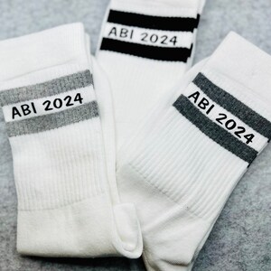 May include: Three white socks with black and grey stripes. Each sock has the text "ABI 2024" printed on the stripe.