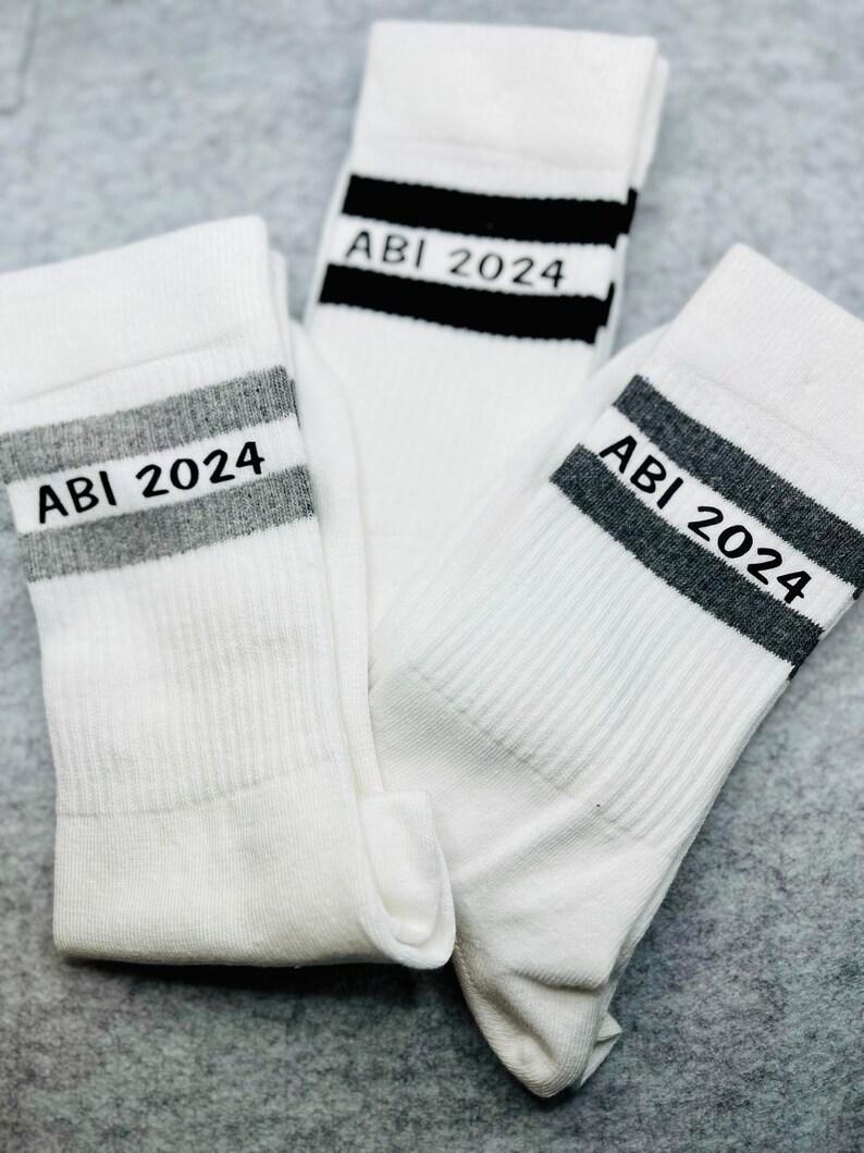 May include: Three white socks with black and grey stripes. Each sock has the text "ABI 2024" printed on the stripe.