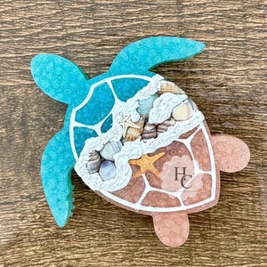 May include: A decorative turtle-shaped object with a turquoise shell and light brown body. The center features a collection of seashells, starfish, and white accents. The object has the letters "HC" on the lower right.