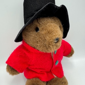 Può includere: Un orsacchiotto marrone con cappello nero e cappotto rosso con bottoni verdi. Il peluche ha un design classico ed è adatto a bambini o collezionisti.