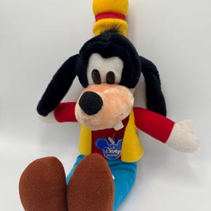 Könnte beinhalten: Ein Plüschtier von Goofy, einer Disney-Figur, in sitzender Position. Das Spielzeug hat einen schwarzen Kopf und Ohren, eine pfirsichfarbene Schnauze und einen gelben Hut mit rotem Band. Er trägt ein rotes Hemd, blaue Hosen und eine gelbe Weste. Das Spielzeug hat ein Disney Original-Etikett.
