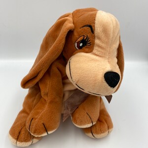May include: A brown and tan plush dog toy with a brown ribbon around its neck. The dog has black eyes and a black nose.