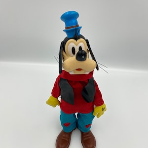 Könnte beinhalten: Ein Vintage-Plüschtier von Goofy, gekleidet in einem roten Hemd, einer schwarzen Weste, einer blauen Jeans und einem blauen Hut. Er hat eine braune Nase und braune Schuhe.