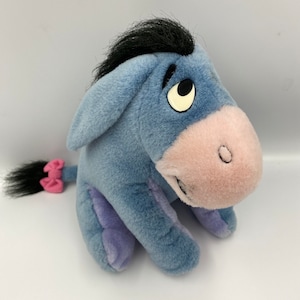 Może przedstawiać: Pluszowy osioł w kolorze niebieskim i fioletowym z różową kokardką na ogonie. Osioł ma czarne włosy i smutny wyraz twarzy.