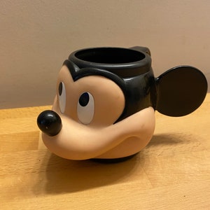 Puede incluir: Una taza de Mickey Mouse de plástico negro y beige con un asa negra y una oreja negra en el lateral.