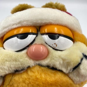 Może przedstawiać: Zbliżenie pluszowej zabawki Garfield w białej czapce Świętego Mikołaja z czerwoną opaską. Pomarańczowy kot ma r&oacute;żowy nos, ż&oacute;łte oczy i ponury wyraz twarzy. Pluszowa zabawka jest wykonana z miękkiego, puszystego materiału.