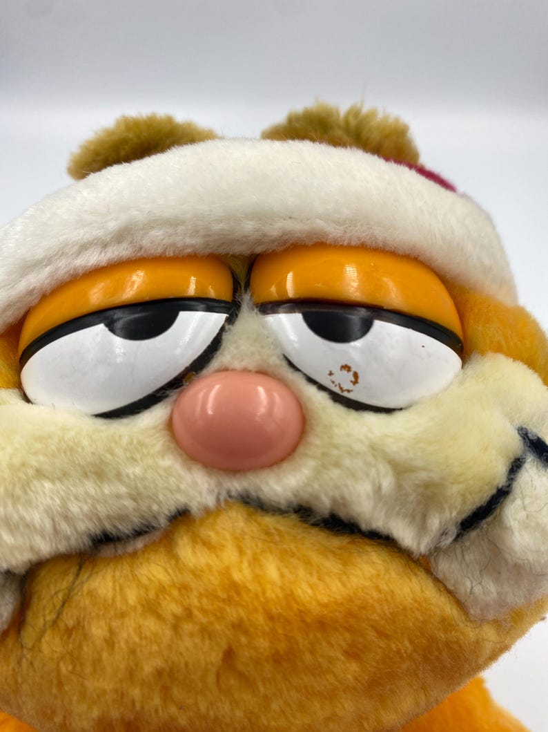 Może przedstawiać: Zbliżenie pluszowej zabawki Garfield w białej czapce Świętego Mikołaja z czerwoną opaską. Pomarańczowy kot ma r&oacute;żowy nos, ż&oacute;łte oczy i ponury wyraz twarzy. Pluszowa zabawka jest wykonana z miękkiego, puszystego materiału.