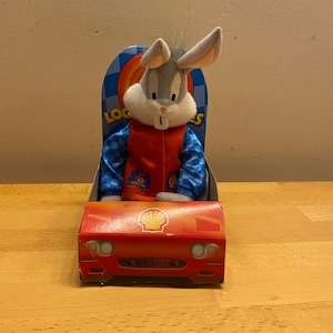 May include: A plush Bugs Bunny toy wearing a blue and white chequered racing jacket and sitting in a red race car with a Shell logo. The car is on a wooden surface.