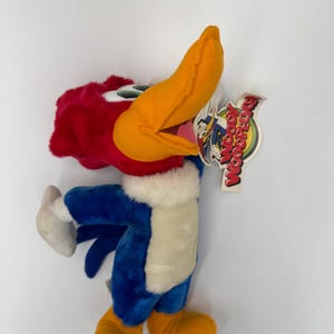 Puede incluir: Un peluche de Woody Woodpecker en una pose dinámica. El juguete presenta una cabeza roja, un pico naranja grande y un cuerpo azul con detalles en blanco. Tiene una etiqueta con el logo de Woody Woodpecker.