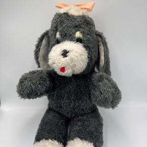 May include: A vintage plush dog toy with grey fur, white accents on the face and paws, and a peach-coloured bow. The dog has large blue eyes and a red mouth. The toy is in a sitting position.