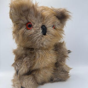 Può includere: Un giocattolo di peluche a forma di koala con pelliccia marrone, occhi rossi e naso nero. Il giocattolo ha un aspetto soffice ed è progettato per assomigliare a un koala. Il koala ha piccoli artigli neri sulle zampe.