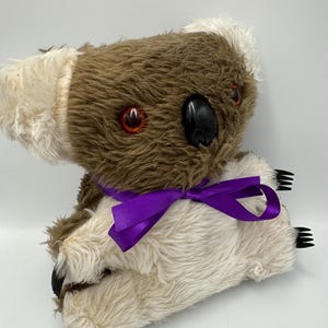 Può includere: Un peluche a forma di koala con pelliccia marrone e crema, con un nastro viola intorno al collo. Il koala ha occhi rossi, un naso nero e artigli neri. Il giocattolo è progettato per assomigliare a un koala.