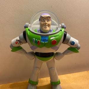 Può includere: Una action figure di plastica verde e bianca di Buzz Lightyear della serie Toy Story. La figura ha un casco viola con una visiera trasparente, una placca toracica verde con le parole "Space Ranger" e un pulsante rosso sul petto. La figura è in piedi con le braccia incrociate e ha un'espressione determinata sul viso.