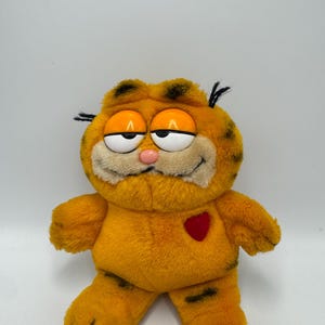 Könnte beinhalten: Ein Garfield-Plüschtier mit orangefarbenem Fell und schwarzen Streifen, großen, halb geschlossenen Kunststoffaugen, einer kleinen rosa Nase und einem roten Herzaufnäher auf der Brust. Die Figur hat ein breites, dezentes Lächeln und schwarze Haarbüschel.