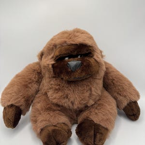 Op de afbeelding: Een gorilla knuffel met zachte, lichtbruine vacht op het lichaam en hoofd, en donkerdere bruine vacht op het gezicht, de handen en de voeten. De gorilla heeft een gladde, donkergrijze driehoekige neus en zichtbare ogen die opzij kijken.