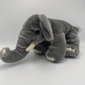 May include: A grey plush elephant toy with a trunk, tusks, and large ears. The stuffed animal has white accents on its feet and trunk. The elephant is lying down on a white surface.