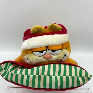 Może przedstawiać: Pluszowa zabawka Garfield w czerwono-białej czapce Świętego Mikołaja, spoczywająca na zielono-białej poduszce w paski z czerwoną obw&oacute;dką. Pomarańczowy kot ma r&oacute;żowy nos i senne oczy.